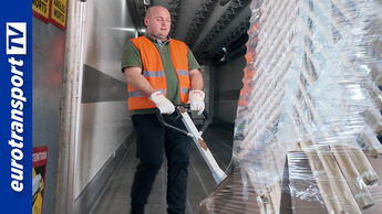 Ein Lkw-Fahrer in Warnweste zieht mit einem Hubwagen eine schwere, in Folie verpackte Ladung im Inneren eines Aufliegers an der Rampe. Die enge Arbeitsumgebung und die körperliche Belastung sind deutlich sichtbar. Links im Bild ist das eurotransportTV-Logo platziert.