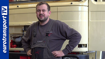 Ein Mann steht in Arbeitskleidung in einer Werkstatt vor einem Lkw, der restauriert wird. Hinter dem Mann das helle Fahrerhaus des Klassikers, links sichtbar das blaue eurotransportTV-Logo.