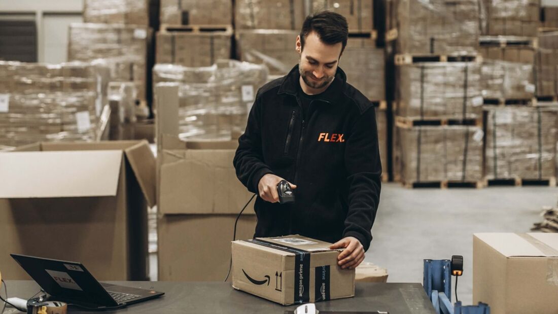 Ein Mitarbeiter des Fulfillment-Dienstleisters Flex scannt ein Amazon-Paket mit einem Handscanner in einem Logistiklager. Im Hintergrund befinden sich Lagerregale und weitere Pakete.