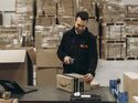 Ein Mitarbeiter des Fulfillment-Dienstleisters Flex scannt ein Amazon-Paket mit einem Handscanner in einem Logistiklager. Im Hintergrund befinden sich Lagerregale und weitere Pakete.