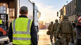 Ein Speditionsmitarbeiter in Warnweste steht vor einem Lkw, während mehrere Bundeswehrsoldaten zu einem Militärfahrzeug gehen. Das Motiv illustriert mögliche Personalausfälle in der Logistik durch den Reservistendienst. (KI-generiertes Symbolbild)
