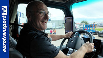Ein lächelnder Lkw-Fahrer sitzt im Cockpit eines modernen Mercedes-Benz eActros und schaut nach draußen. Im Hintergrund sind das Testgelände und mehrere Fahrzeuge zu sehen. Links befindet sich das eurotransportTV-Logo.