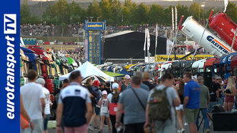 Eine Menschenmenge läuft beim Truck Grand Prix über das Eventgelände, flankiert von langen Reihen parkender Lkw auf beiden Seiten; im Hintergrund sind Zuschauertribünen, eine Bühne und ein großer Goodyear-Anzeigeturm zu sehen; am linken Bildrand befindet sich das vertikale Logo von eurotransportTV.