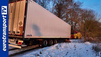Festgefahrener weißer Kühlauflieger von Schmitz Cargobull steht bei Schnee und Eis schräg in der Bankette, im Hintergrund ein gelbes Bergefahrzeug im Einsatz, links das eurotransportTV-Logo.