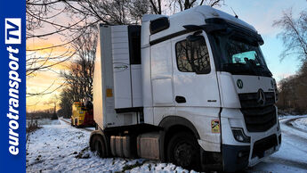 Festgefahrener weißer Mercedes-Benz Actros mit Kühlauflieger bei Schnee und Eis, im Hintergrund ein Abschleppfahrzeug im Einsatz, eurotransportTV-Logo links im Bild.