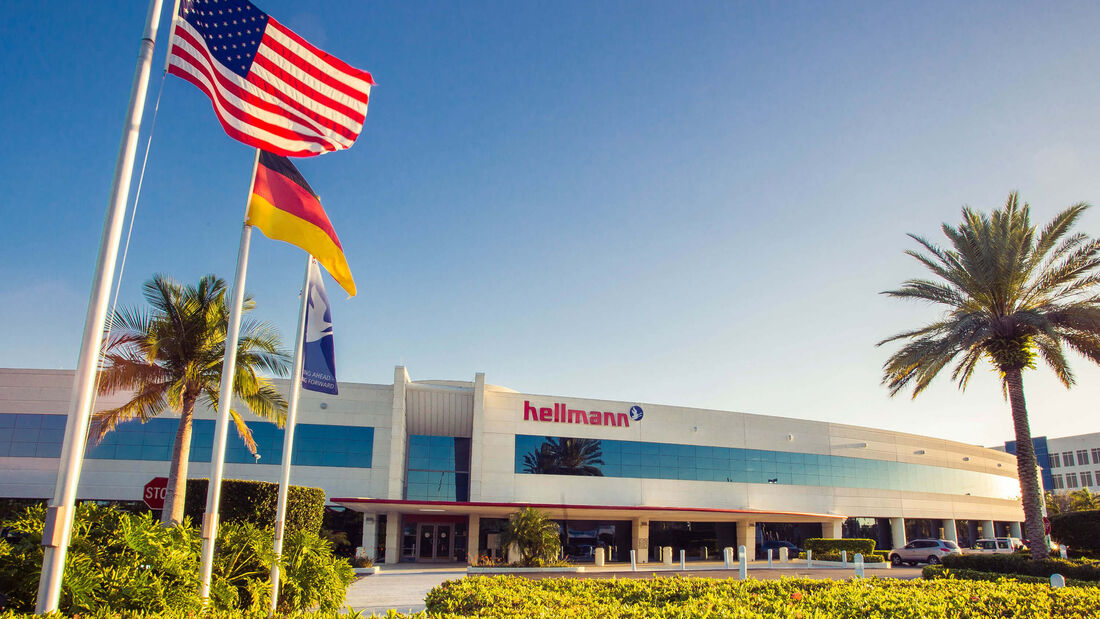 Hellmann-Standort in Miami, Florida, mit drei Flaggen im Vordergrund: der deutschen Nationalflagge, der US-amerikanischen Flagge und der Unternehmensflagge von Hellmann Worldwide Logistics.