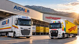 KI-generiertes Bild auf Basis von Unternehmensfotos: Ein weißer Aigner-Sattelzug und ein gelb-weißer Nothegger-Lkw stehen vor einer Logistikhalle. Im Hintergrund sind weitere Lkw und eine Berglandschaft zu sehen.
