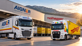 KI-generiertes Bild auf Basis von Unternehmensfotos: Ein weißer Aigner-Sattelzug und ein gelb-weißer Nothegger-Lkw stehen vor einer Logistikhalle. Im Hintergrund sind weitere Lkw und eine Berglandschaft zu sehen.