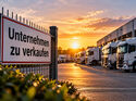 KI-generiertes Symbolbild eines Speditionshofs mit mehreren geparkten Lkw vor einer Logistikhalle. Im Vordergrund hängt ein Schild mit der Aufschrift „Unternehmen zu verkaufen“ am Zaun.