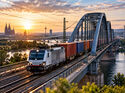 KI-generiertes Symbolbild eines elektrischen Güterzugs mit Containern auf einer großen Eisenbahnbrücke über einem Fluss. Im Hintergrund ist eine europäische Großstadt bei Sonnenuntergang zu sehen.