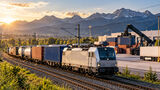 KI-generiertes Symbolbild eines elektrischen Güterzugs mit Containern vor einer Alpenkulisse. Neben den Gleisen befindet sich ein Umschlagterminal mit Containerstapeln, einem Reachstacker und einer Logistikhalle.