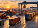 KI-generiertes Symbolbild eines großen Containerterminals im Sonnenuntergang. Ein Sattelzug transportiert einen Seecontainer über das Terminalgelände, während ein Containerzug auf den Gleisen steht und Portalkräne Container umschlagen.