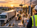 KI-generiertes Symbolbild eines modernen Logistikzentrums bei Sonnenuntergang. Lkw, Gabelstapler und Fördertechnik prägen den Warenumschlag, während ein Mitarbeiter mit Tablet die Abläufe überwacht.