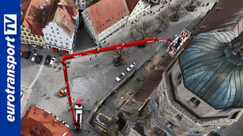 Luftaufnahme einer historischen Altstadt mit engem Straßennetz, in der ein roter Lkw mit ausgefahrener 100 Meter Arbeitsbühne bis an das Dach eines Kirchturms reicht, während mehrere Personen im Korb arbeiten; das eurotransportTV-Logo ist am linken Bildrand sichtbar.