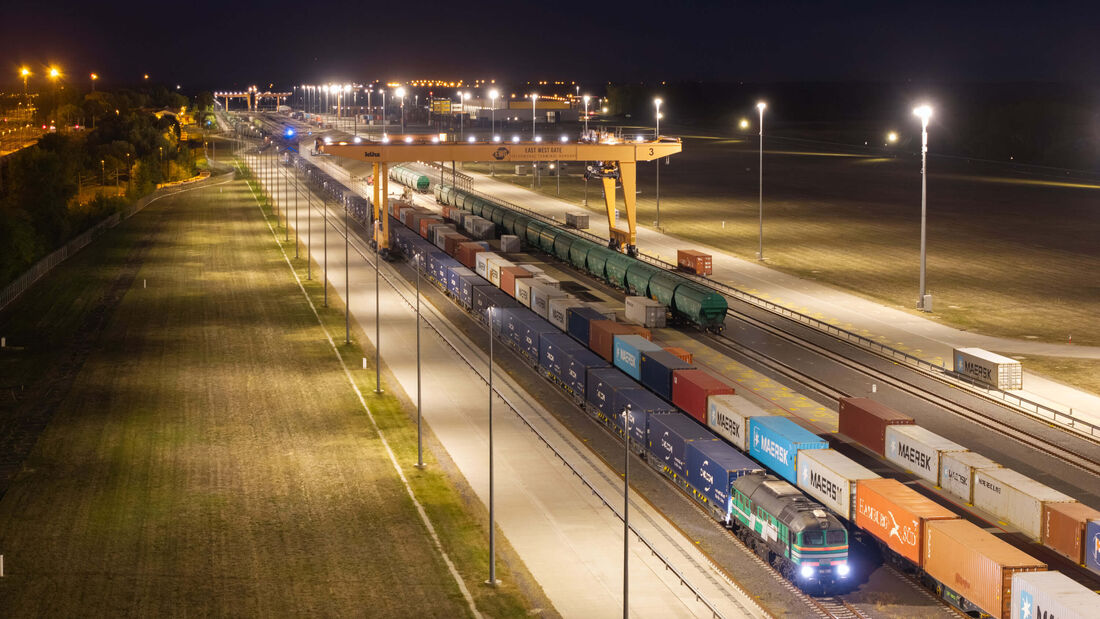 Nächtliche Luftaufnahme des Intermodalterminals East-West Gate im ungarischen Fényeslitke. Ein Containerzug steht unter einem großen gelben Portalkran. Auf den Gleisen und Umschlagflächen sind zahlreiche Container zu sehen, darunter mehrere von CMA CGM und Maersk.