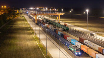 Nächtliche Luftaufnahme des Intermodalterminals East-West Gate im ungarischen Fényeslitke. Ein Containerzug steht unter einem großen gelben Portalkran. Auf den Gleisen und Umschlagflächen sind zahlreiche Container zu sehen, darunter mehrere von CMA CGM und Maersk.