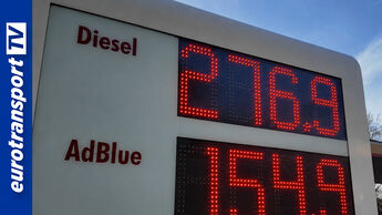 Preisanzeige an einer Tankstelle mit stark gestiegenem Dieselpreis von 2,76 Euro pro Liter und AdBlue-Preis darunter, aufgenommen aus leichter Untersicht bei Tageslicht. Im Hintergrund sind Teile der Tankstelle und Umgebung zu erkennen. Links im Bild befindet sich ein blauer Balken mit dem eurotransportTV-Logo.