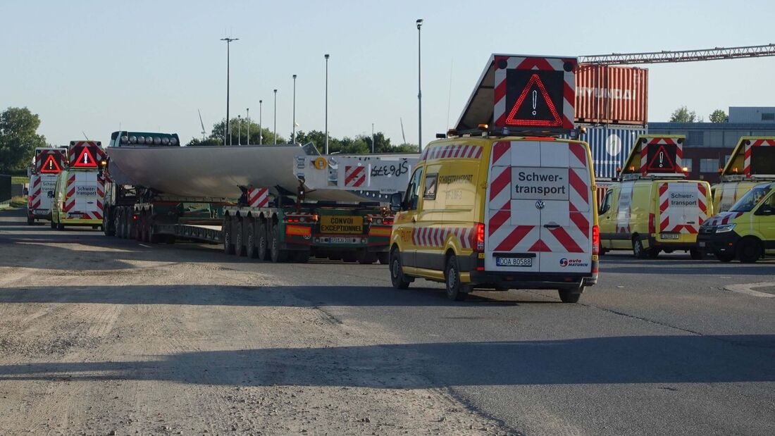 Schwertransport mit einem großen Industriebauteil auf einem Tieflader, begleitet von mehreren gelben Sicherungsfahrzeugen mit Warnleuchten und Schwertransport-Kennzeichnung.