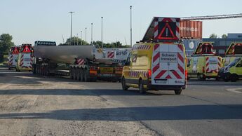 Schwertransport mit einem großen Industriebauteil auf einem Tieflader, begleitet von mehreren gelben Sicherungsfahrzeugen mit Warnleuchten und Schwertransport-Kennzeichnung.