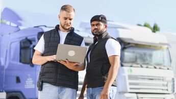 Truckers collaborating on logistics management using laptop. Two male truckers discussing transport routes and fleet details using a laptop at a truck parking lot, ensuring efficient delivery
