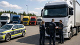 Zollbeamte kontrollieren einen Lkw-Fahrer und seine Unterlagen auf einem Betriebshof mit mehreren Lastwagen.