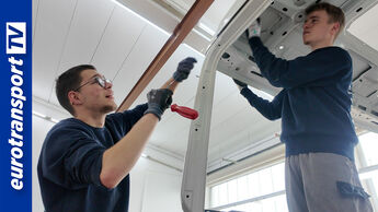 Zwei Auszubildende arbeiten in einer hellen Lkw-Werkstatt an der Kabine eines MAN TGA und führen Karosseriearbeiten durch, eurotransportTV-Logo links im Bild sichtbar.