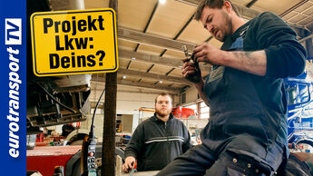 Zwei Lkw-Mechaniker bei der Arbeit in einer Werkstatt, einer sitzt mit schmutzigen Händen auf einem Lkw-Rad und prüft ein Teil. Links oben im Bild ist ein gelbes Schild mit der Aufschrift „Projekt Lkw: Deins?“ zu sehen. Am linken Rand befindet sich das eurotransportTV-Logo.