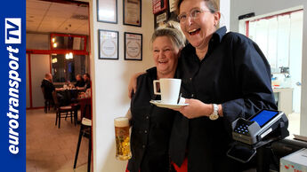 Zwei Mitarbeiterinnen des Hoyer Autohofs Rade lachen herzlich im Gastraum, eine hält ein Bier, die andere eine Kaffeetasse. Im Hintergrund sitzen Gäste an Tischen. Links ist das eurotransportTV-Logo sichtbar.