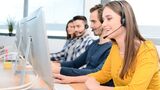 portrait of beautiful and cheerful young woman telephone operator with headset working on desktop computer in row in a customer service call support helpline business center with teamworker in background